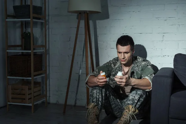 Militar deprimido sosteniendo pastillas mientras está sentado en el suelo por la noche - foto de stock