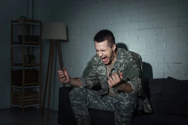 Soldado enojado con trastorno de estrés postraumático gritando en el sofá en casa por la noche - foto de stock
