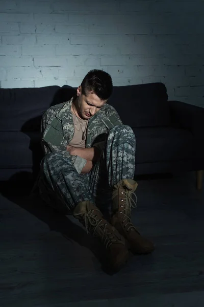 Militaire déprimé en uniforme militaire souffrant d'un trouble de stress post-traumatique et assis sur le sol dans une pièce sombre — Photo de stock