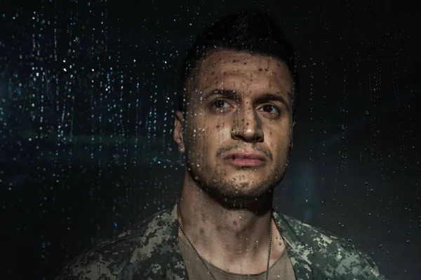 Sad serviceman in military uniform looking through rainy window while suffering from post traumatic stress disorder — Stock Photo