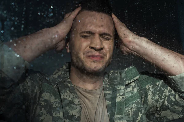 Hombre de servicio ansioso en uniforme militar que sufre de trastorno de estrés postraumático mientras está de pie junto a la ventana con gotas de lluvia - foto de stock