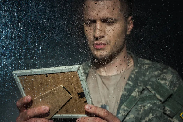 Сумний військовослужбовець дивиться на фоторамку і має спалахи, стоячи поруч з вікном з краплями дощу — стокове фото
