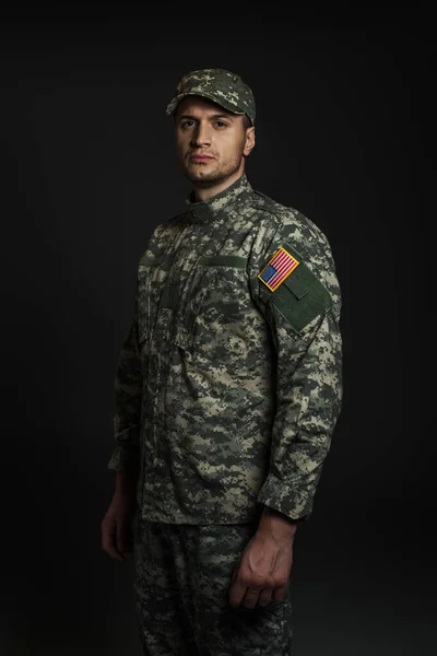 Soldado americano patriótico en uniforme y gorra mirando a la cámara aislada en negro - foto de stock