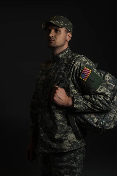 Patriotic soldier in uniform with American flag holding backpack isolated on black — Stock Photo