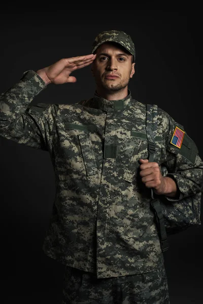 Soldado patriótico en uniforme con bandera americana saludo aislado en negro - foto de stock