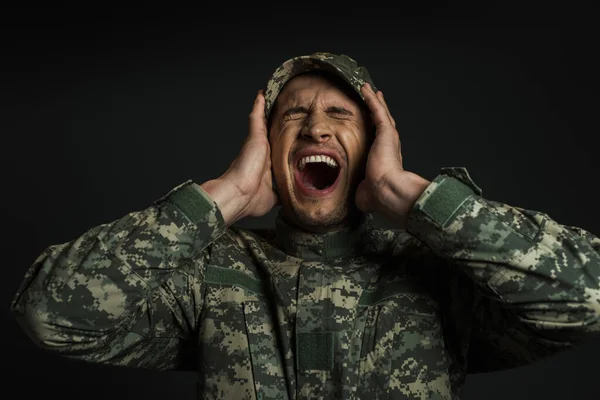 Soldado estresado en uniforme de camuflaje y gorra gritando mientras sufría de ptsd aislado en negro - foto de stock