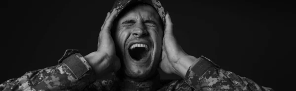 Monochrome photo of stressed soldier in uniform and cap screaming while suffering from ptsd isolated on black, banner — Stock Photo