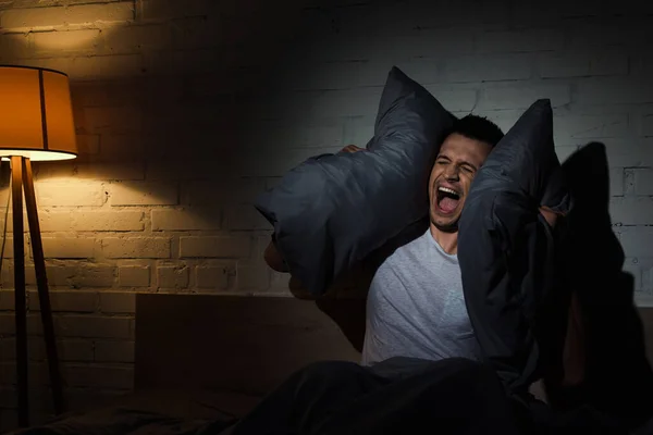 Scared young man screaming while having nightmares and covering ears with pillows at night — Stock Photo