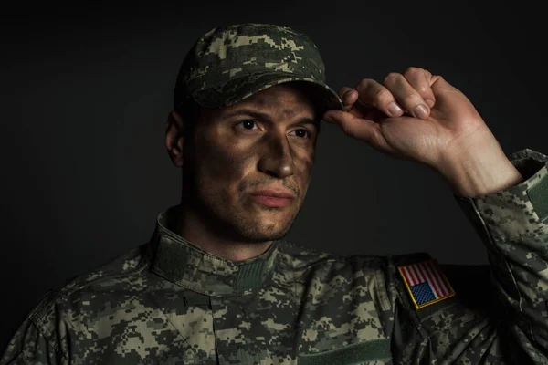 Military man with dirt on face standing in uniform and adjusting cap isolated on grey — Stock Photo