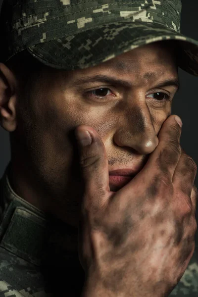 Soldado estresado con suciedad en la cara tocando la cara y mirando hacia otro lado mientras sufre de ptsd aislado en gris - foto de stock