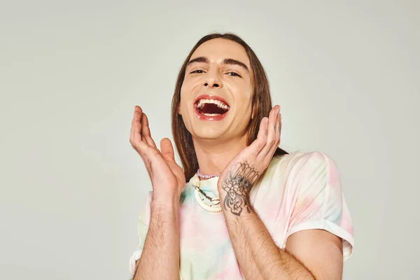 Hombre gay joven tatuado y excitado con el pelo largo y la boca abierta mirando a la cámara y el gesto con las manos en el fondo gris, orgullo concepto de mes - foto de stock