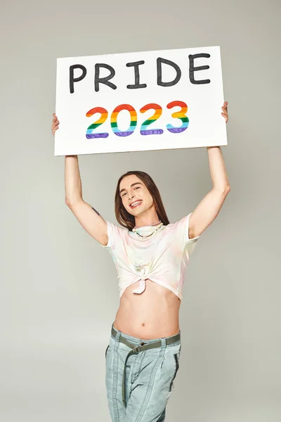 Cheerful young gay man with tattoo and long hair standing in denim jeans and tied knot on t-shirt showing his belly and holding pride 2023 placard on grey background — Stock Photo