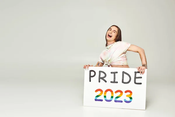 Возбужденный молодой гей-активист с татуировкой и длинными волосами стоя с открытым ртом и держа гордость 2023 плакат на сером фоне, lgbt сообщества праздник в июне — стоковое фото