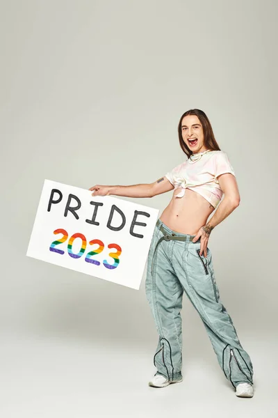Cheerful young gay man with tattoo, long hair and open mouth standing in denim jeans and tied knot on t-shirt showing his belly while holding pride 2023 placard on grey background — Stock Photo
