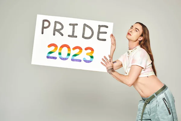 Молодой гей с татуировкой и длинными волосами, стоящий в джинсах в джинсах и завязанный узел на футболке, показывающий свой живот, держа плакат гордости 2023 года на сером фоне — стоковое фото