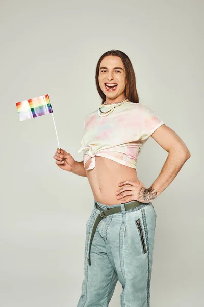 Cheerful gay man with tattoo and long hair standing with hand on hip and bare belly while holding rainbow flag for pride month on grey background — Stock Photo