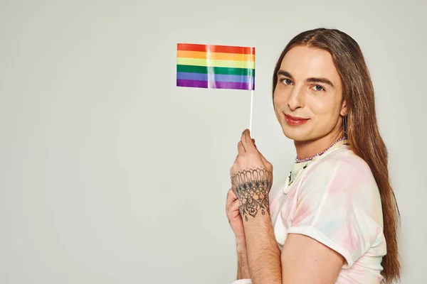 Весёлый гей с татуировкой на руке и длинными волосами, держащий радужный флаг для гордости месяц и улыбающийся, глядя на камеру, изолированную на сером фоне — стоковое фото