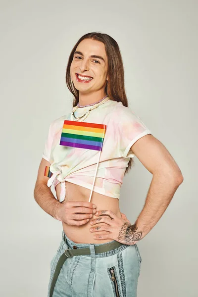 Allegro gay uomo con tatuaggio e lunghi capelli in piedi con mano su anca e ventre nudo mentre tenendo arcobaleno lgbt bandiera per orgoglio mese su sfondo grigio — Foto stock