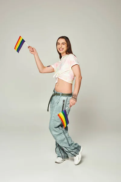 Cheerful and tattooed gay man with long hair and bare belly standing in denim jeans while holding rainbow flags for pride month on grey background — Stock Photo