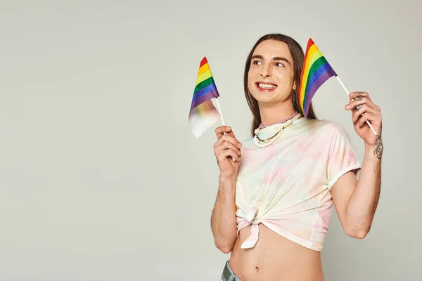 Happy and tattooed homosexual man with long hair and bare belly smiling while holding rainbow lgbt flags for pride month on grey background — Stock Photo