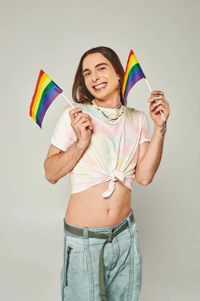 Happy and tattooed gay man with long hair and bare belly standing in denim jeans while holding rainbow flags for pride month on grey background — Stock Photo