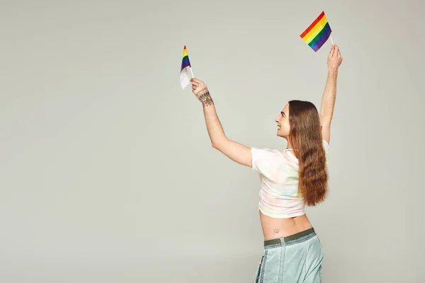 Cheerful and tattooed gay man with long hair standing in denim jeans while holding rainbow flags for pride month above head on grey background — Stock Photo