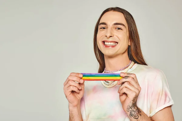 Happy and tattooed gay man with long hair and tie dye t-shirt holding rainbow lgbt flag for pride month and smiling while looking at camera on grey background — Stock Photo