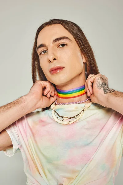 Tatuato gay uomo con lungo capelli e cravatta tintura t-shirt tenendo arcobaleno lgbt bandiera vicino collo durante orgoglio mese e guardando fotocamera su sfondo grigio — Foto stock