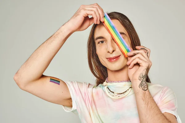Feliz y tatuado hombre gay con el pelo largo y corbata tinte camiseta sosteniendo arco iris lgbt bandera cerca de la cara durante el mes de orgullo y sonriendo mientras mira a la cámara en el fondo gris - foto de stock