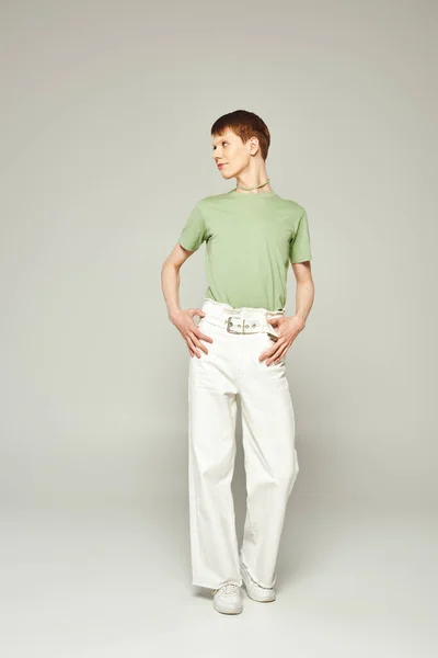 In voller Länge junger queerer Mensch mit glänzendem Lipgloss, der in grünem T-Shirt und weißen Jeans steht, während er während eines stolzen Monats auf grauem Hintergrund wegschaut — Stockfoto