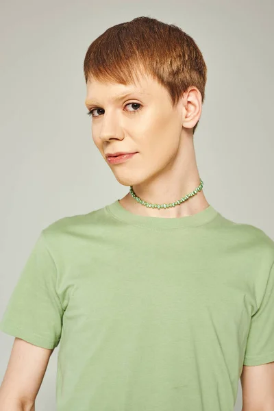 Portrait of young nonbinary person with shiny lip gloss standing in green t-shirt and looking at camera during pride month on grey background — Stock Photo