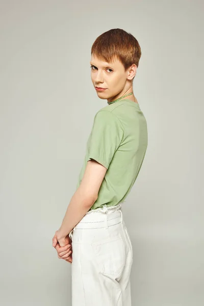 Junge queere Person mit Lipgloss in weißen Jeans und grünem T-Shirt, während sie im Monat Stolz auf grauen Hintergrund in die Kamera schaut — Stockfoto