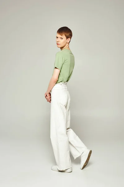 Полная длина молодого странного человека с блеском для губ, стоящего в зеленой футболке и джинсах в белых джинсах, глядя в камеру на месяц гордости на сером фоне — стоковое фото