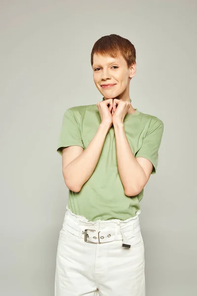 Happy queer person with shiny lip gloss standing in white denim jeans and green t-shirt while looking at camera and holding hands in heart shape during pride month on grey background — Stock Photo