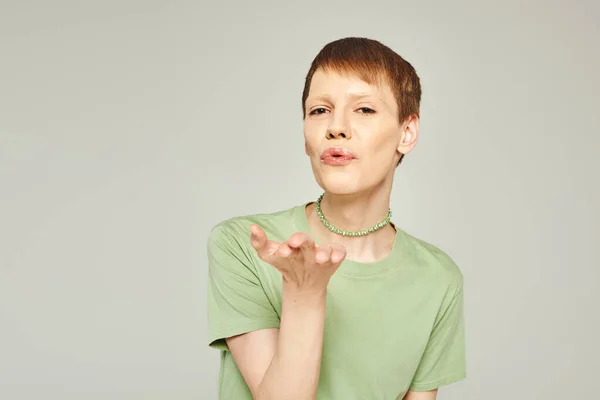 Jovem pessoa não-binária com brilho labial em pé em t-shirt verde e soprando beijo de ar enquanto olha para a câmera durante o mês de orgulho no fundo cinza — Fotografia de Stock