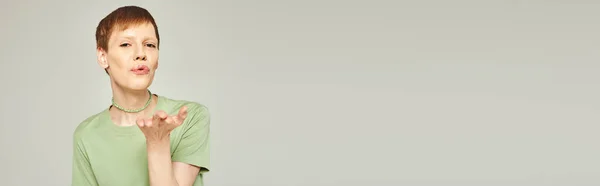 Jovem queer pessoa com brilho labial de pé em t-shirt verde e soprando beijo de ar enquanto olha para a câmera durante o mês de orgulho no fundo cinza, banner — Fotografia de Stock
