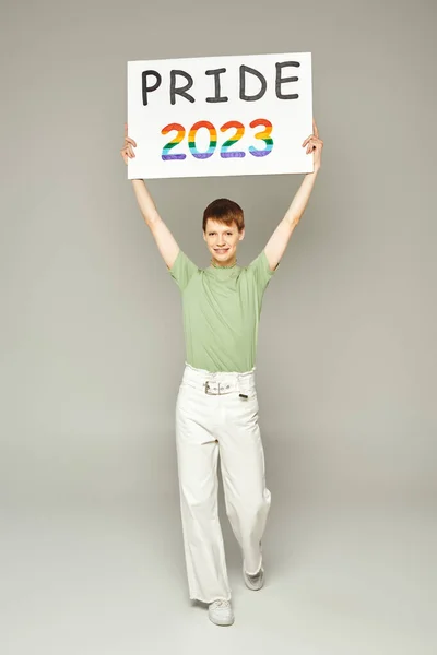 Full length of happy young queer person with lip gloss standing in white denim jeans and green t-shirt while holding pride 2023 placard on grey background — Stock Photo