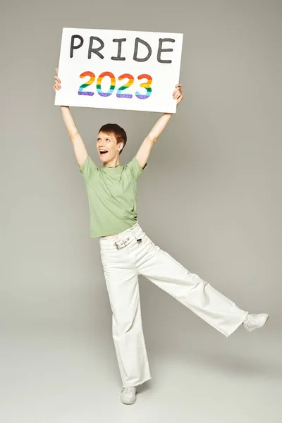 Полная длина веселый странный человек с блеском для губ и открытым ртом стоя в белых джинсах джинсы джинсы и зеленая футболка, держа гордость 2023 плакат на сером фоне — стоковое фото