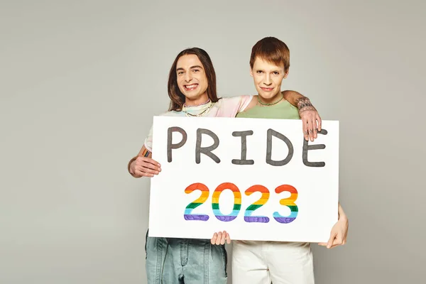 Cheerful lgbt friends holding pride 2023 placard and looking at camera while celebrating lgbtq community holiday in June on grey background in studio — Stock Photo