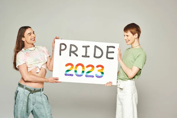 Fröhliche schwule Männer mit stolzem 2023-Plakat, während sie im Juni den lgbtq-Community-Feiertag feiern und gemeinsam auf grauem Hintergrund im Studio stehen — Stockfoto