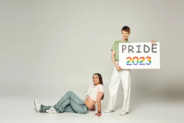 Happy gay man holding pride 2023 placard while standing next to smiling queer friend with bare belly and celebrating lgbtq community holiday in June on grey background in studio — Stock Photo