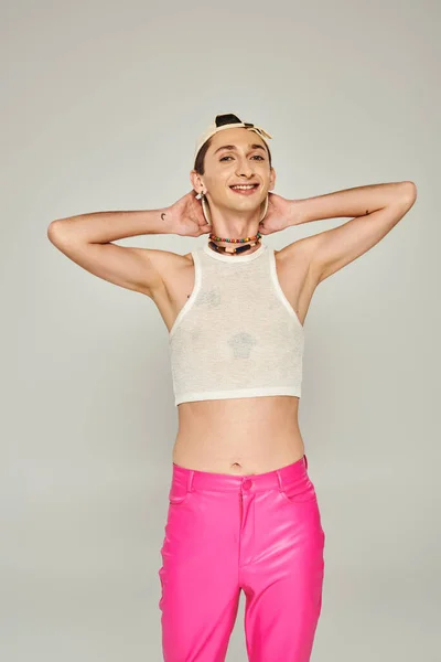 Счастливый и татуированный молодой гей в бейсболке, топ и розовые брюки улыбаются, позируя на сером фоне, концепция Дня гордости — стоковое фото