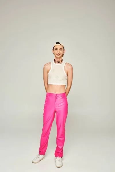 Full length of happy and tattooed gay man in baseball cap, crop top and pink pants smiling while posing on grey background, pride day concept — Stock Photo