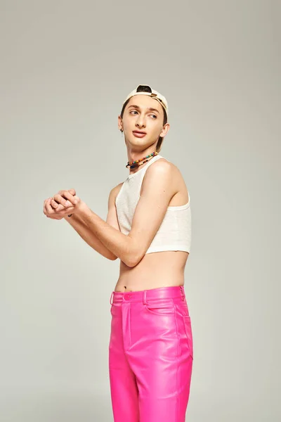 Hombre gay joven tatuado en gorra de béisbol, top de la cosecha, vientre desnudo y pantalones de color rosa posando con las manos apretadas y mirando hacia otro lado sobre fondo gris, concepto del día del orgullo - foto de stock