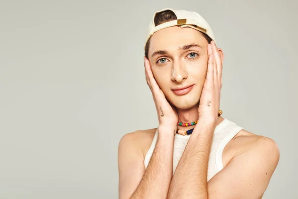 Portrait of tattooed young gay man with blue eyes posing in baseball cap and colorful beads and looking at camera on grey background, pride day concept — Stock Photo