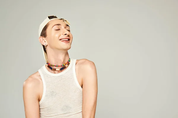 Portrait of pleased young gay man with closed eyes posing in baseball cap, tank top and colorful beads on grey background, pride day concept — Stock Photo