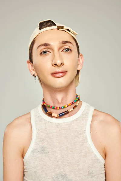 Portrait of young gay man with blue eyes eyes posing in baseball cap, white tank top and colorful beads on grey background, pride day concept — Stock Photo