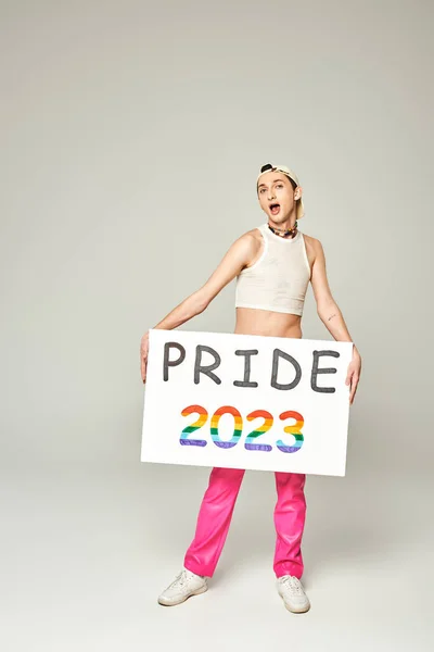 Tätowierter junger schwuler Mann mit Baseballkappe, bauchfreiem Oberteil und pinkfarbener Hose mit stolzem 2023-Plakat, während er mit geöffnetem Mund vor grauem Hintergrund in die Kamera blickt — Stockfoto
