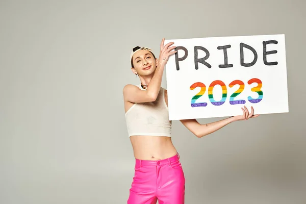 Tattooed and pleased young gay man in baseball cap, crop top, and pink pants holding pride 2023 placard while standing and looking at camera on grey background — Stock Photo