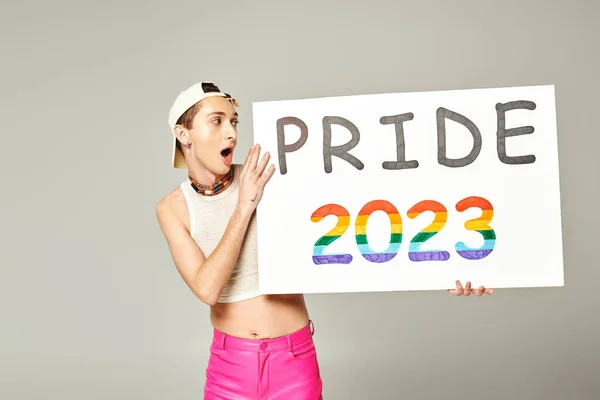 Татуйований і шокований гей чоловік у бейсбольній шапці, верхній частині врожаю, і рожеві штани, що тримають гордість плакату 2023 року, стоячи з відкритим ротом на сірому фоні — стокове фото